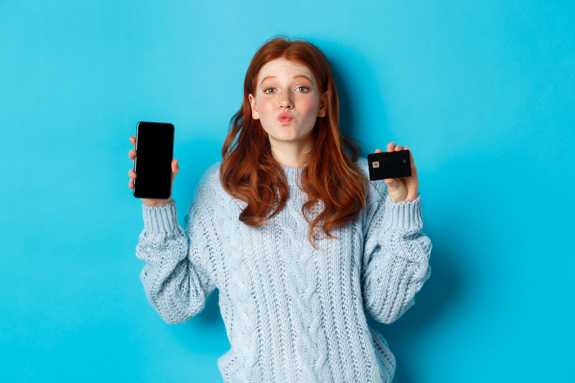 Excited redhead girl showing mobile phone screen and credit card, demonstrating online store or